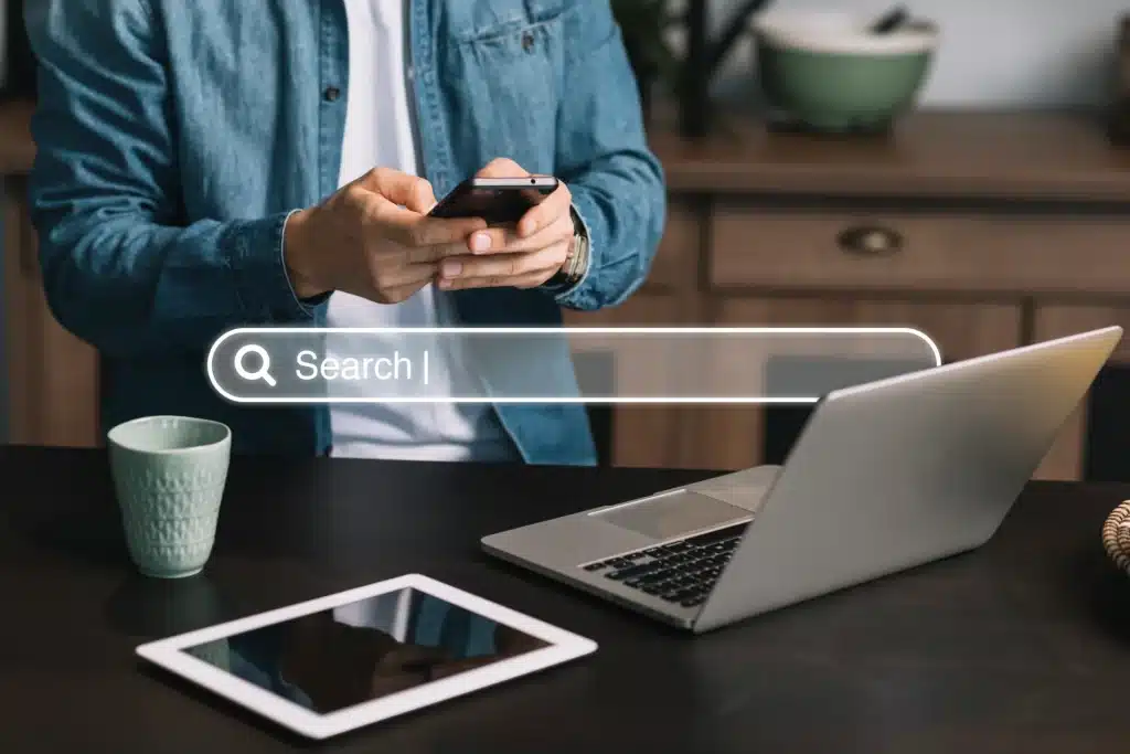 Eine Person in einer Jeansjacke nutzt ein Smartphone an einem Schreibtisch mit Laptop, Tablet und grüner Tasse.