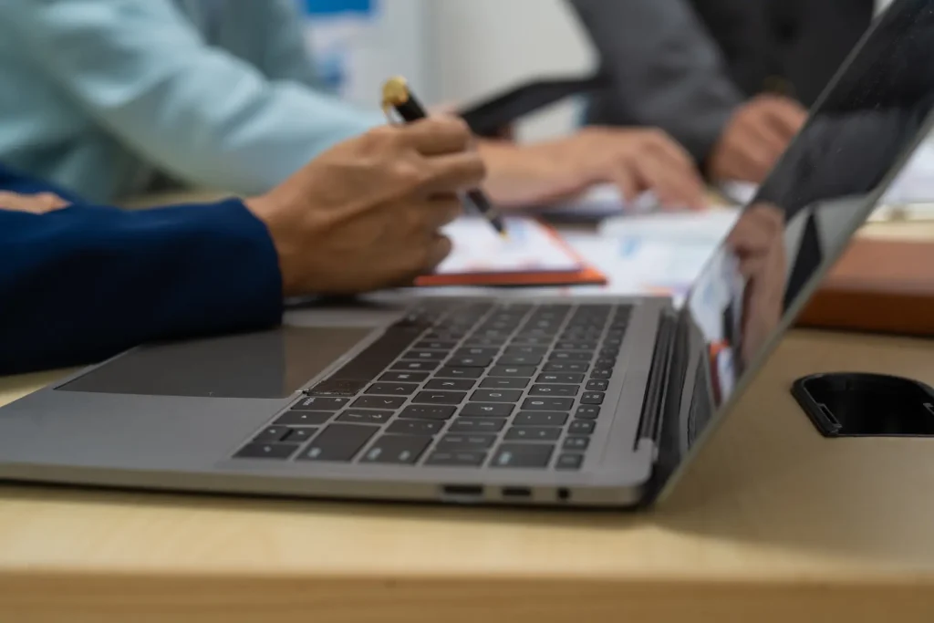Eine Nahaufnahme eines Laptops auf einem Schreibtisch. Zwei Personen mit Stiften diskutieren über Strategien.